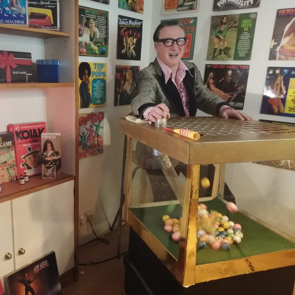 Comedy Bingo Host Peter Perky sitting by his retro 1970s blower bingo machine